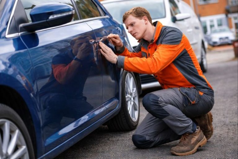 Automotive locksmith technician wearing an orange and gray uniform unlocking a blue car door, professional car lockout service without damage.
